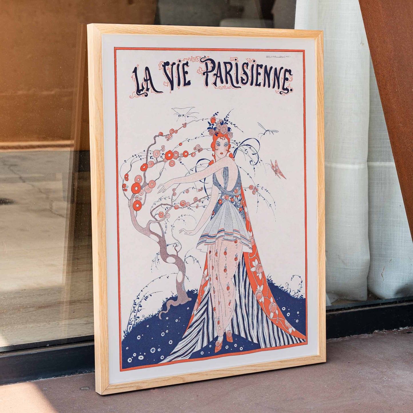 Elegant Art Deco poster of a woman in a floral dress standing by a blossoming tree La Vie Parisienne by Umberto Brunelleschi, framed and displayed in home decor